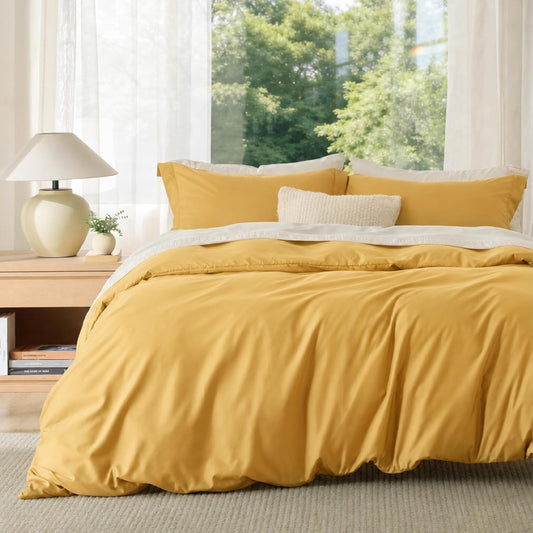A mustard yellow bedding set, including a duvet cover and matching pillow shams, styled on a bed in a bright bedroom with a wooden nightstand, lamp, and a large window showing green trees outside.