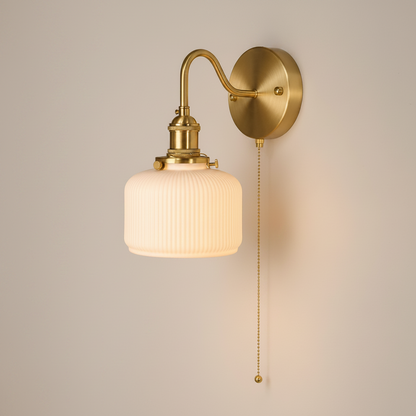 A wall-mounted sconce with a ribbed glass shade, shown lit in a real interior hallway. The fixture has a warm brass/gold finish and a pull chain.