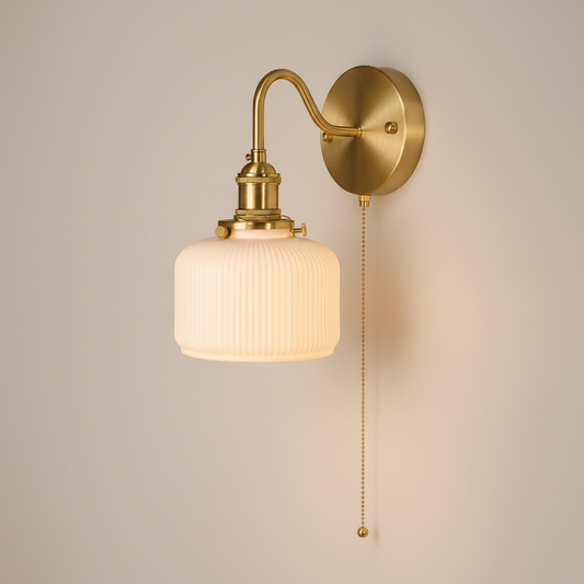 A wall-mounted sconce with a ribbed glass shade, shown lit in a real interior hallway. The fixture has a warm brass/gold finish and a pull chain.