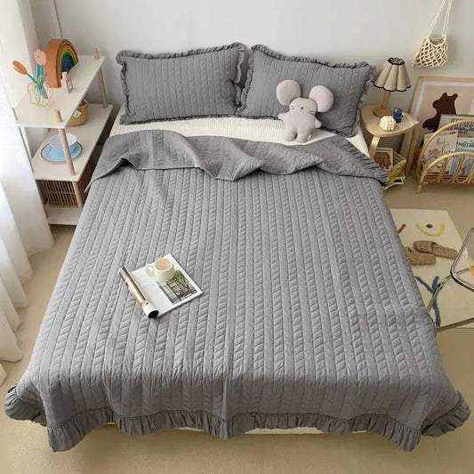 Gray textured bedspread on a bed in a bedroom setting with pillows and a teddy bear.