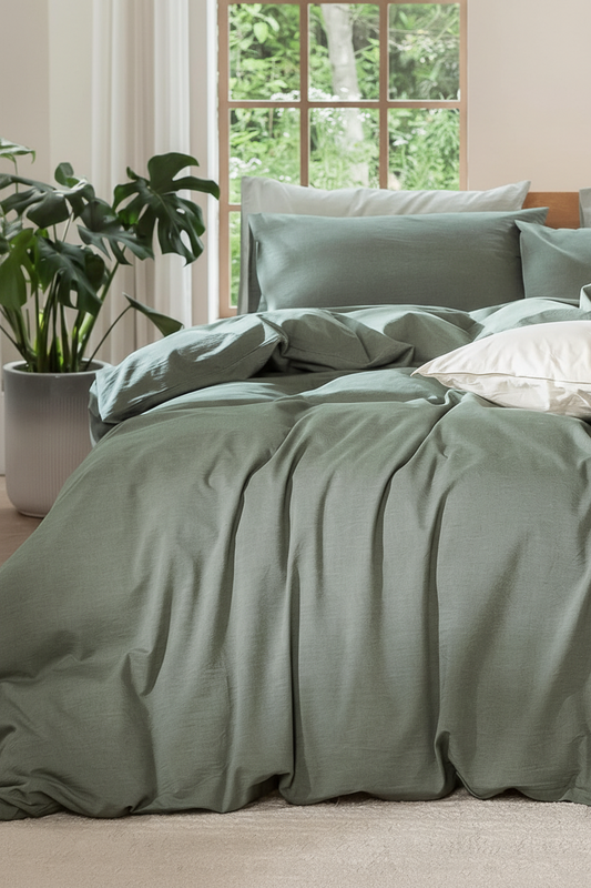 A bed dressed in a sage green bedding set with matching pillows and a rumpled duvet, styled in a bright bedroom with a window, curtains, and a potted plant.