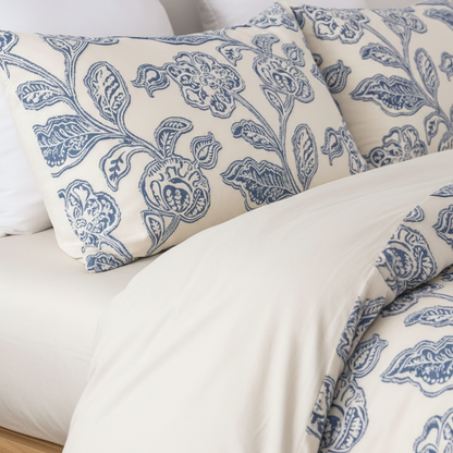 A close-up of a bed dressed in white bedding with two decorative pillows and a duvet featuring a blue floral and botanical pattern.