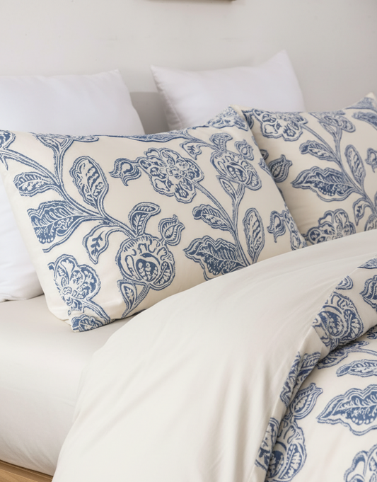 A close-up of a bed dressed in white bedding with two decorative pillows and a duvet featuring a blue floral and botanical pattern.