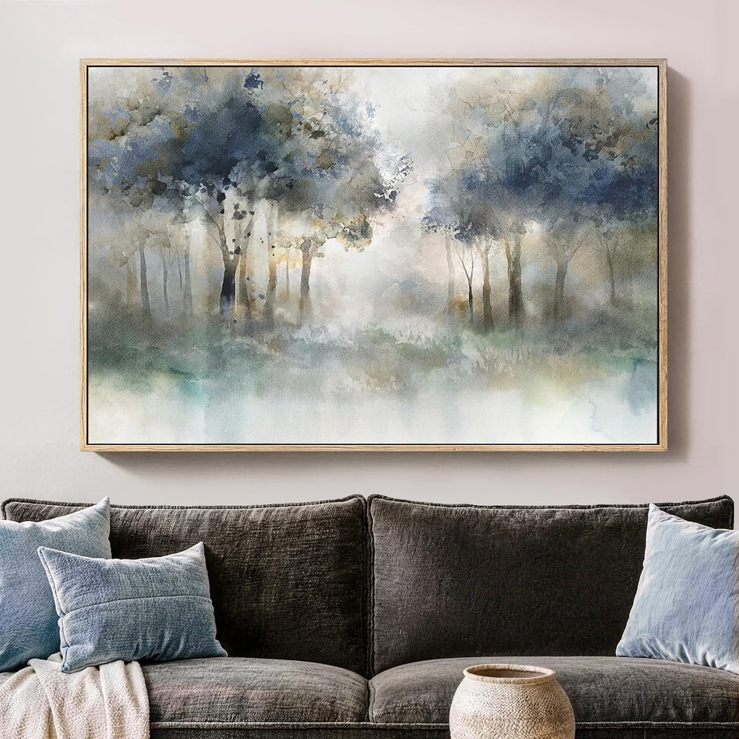 A framed watercolor painting of a misty forest with blue, gray, and white tones, displayed on a light beige wall above a dark gray sofa with blue pillows.