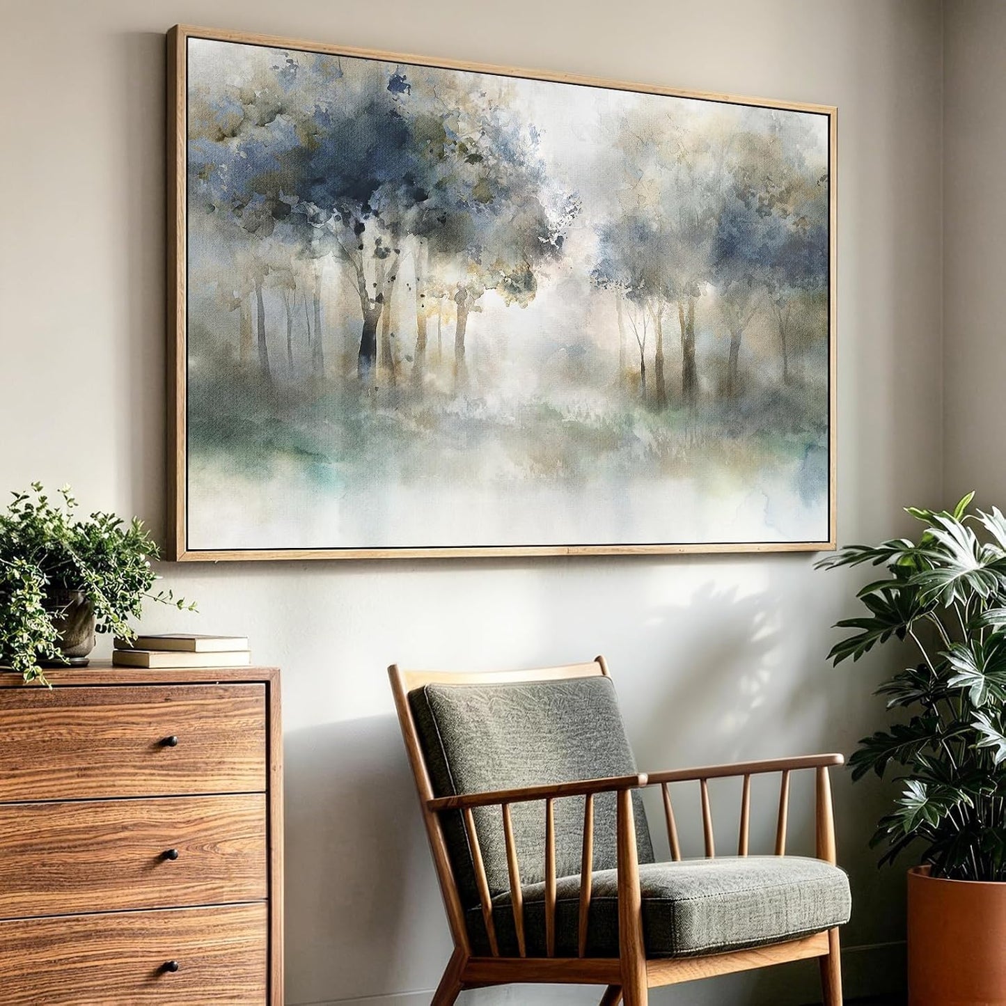 A large framed landscape painting of a misty blue-gray forest hangs on a beige wall above a mid-century modern wooden armchair, with a wooden chest and potted plants nearby.