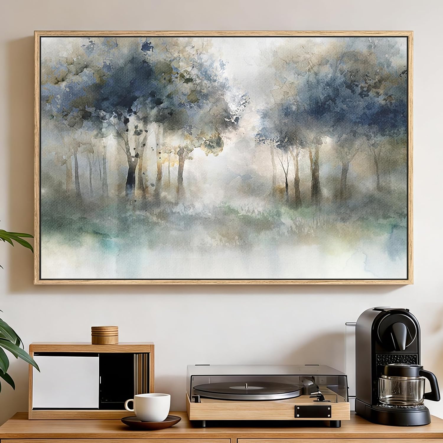 A framed watercolor landscape painting of a misty forest with blue-gray trees and soft green undertones, displayed on a beige wall above a wooden console table with a record player, coffee maker, and cup.
