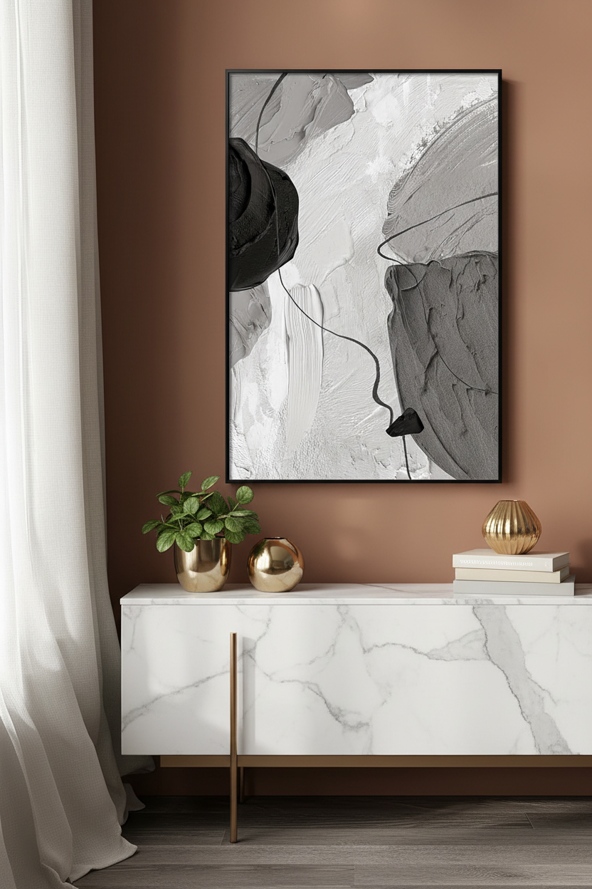 A framed abstract black, white, and gray wall art print hangs on a warm brown wall above a white marble console table with gold accents, styled with a small plant and decorative spheres.