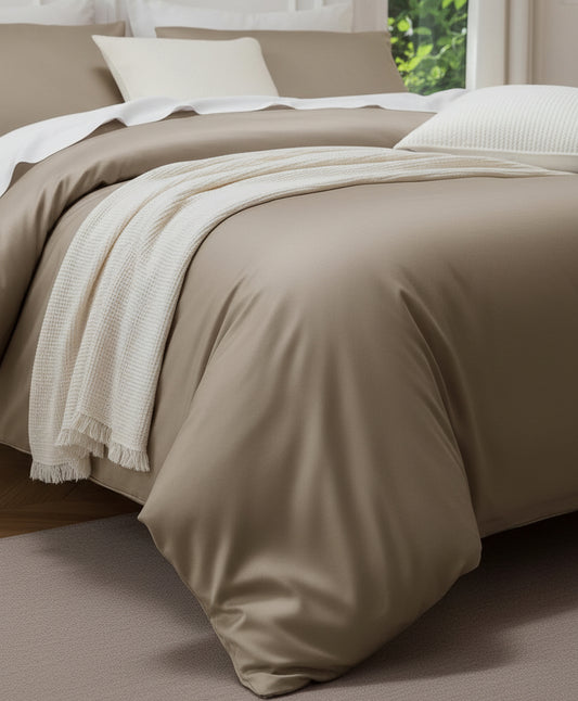 A neatly made bed in a styled bedroom setting, featuring a champagne beige satin duvet cover, matching pillow shams, white accent pillows, and a cream knit throw blanket draped over the foot of the bed. The background includes a white paneled wall, a window with green foliage outside, and bedside tables with lamps.