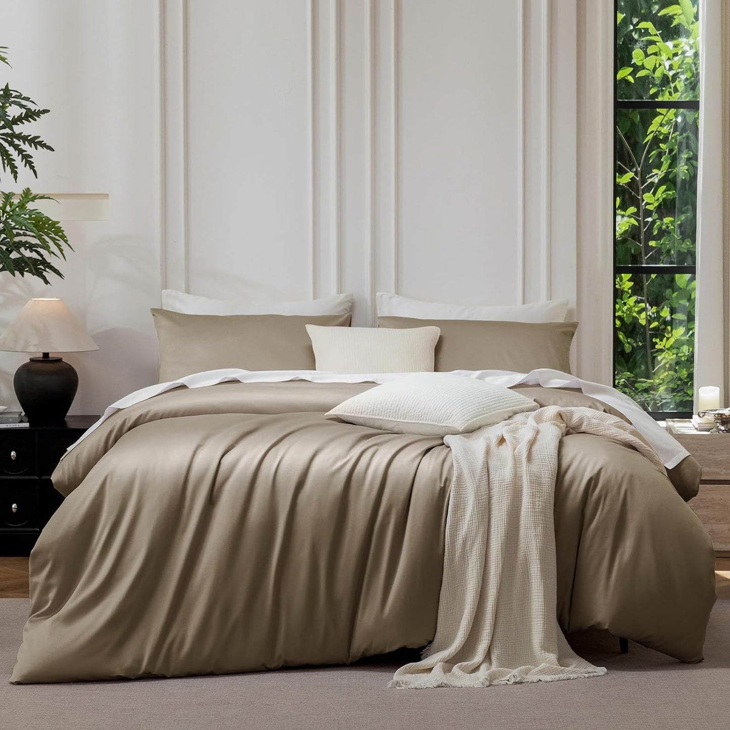 A neatly made bed in a styled bedroom setting, featuring a champagne beige satin duvet cover, matching pillow shams, white accent pillows, and a cream knit throw blanket draped over the foot of the bed. The background includes a white paneled wall, a window with green foliage outside, and bedside tables with lamps.