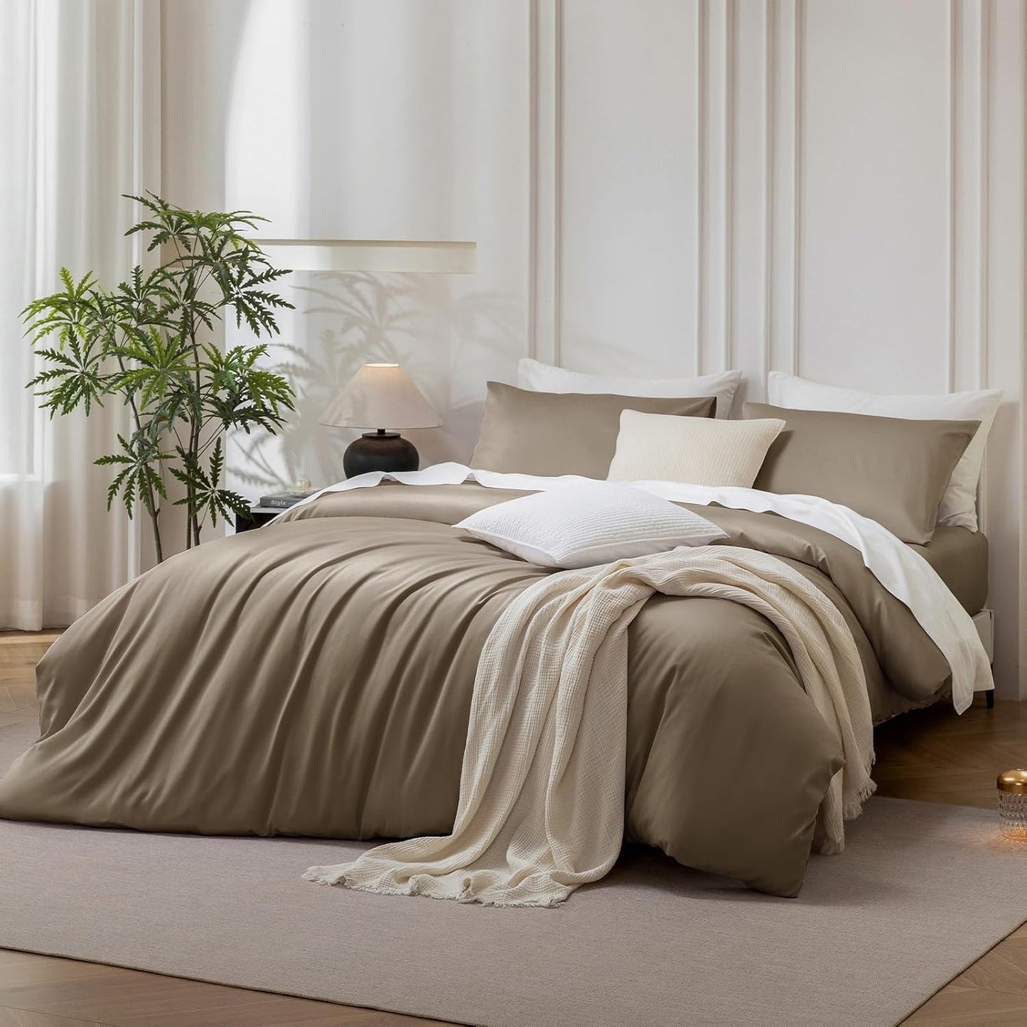 A neatly made bed in a softly lit bedroom, dressed in taupe and cream bedding with white pillows and a cream throw blanket. The room has paneled walls, a plant, a bedside lamp, and a light rug.