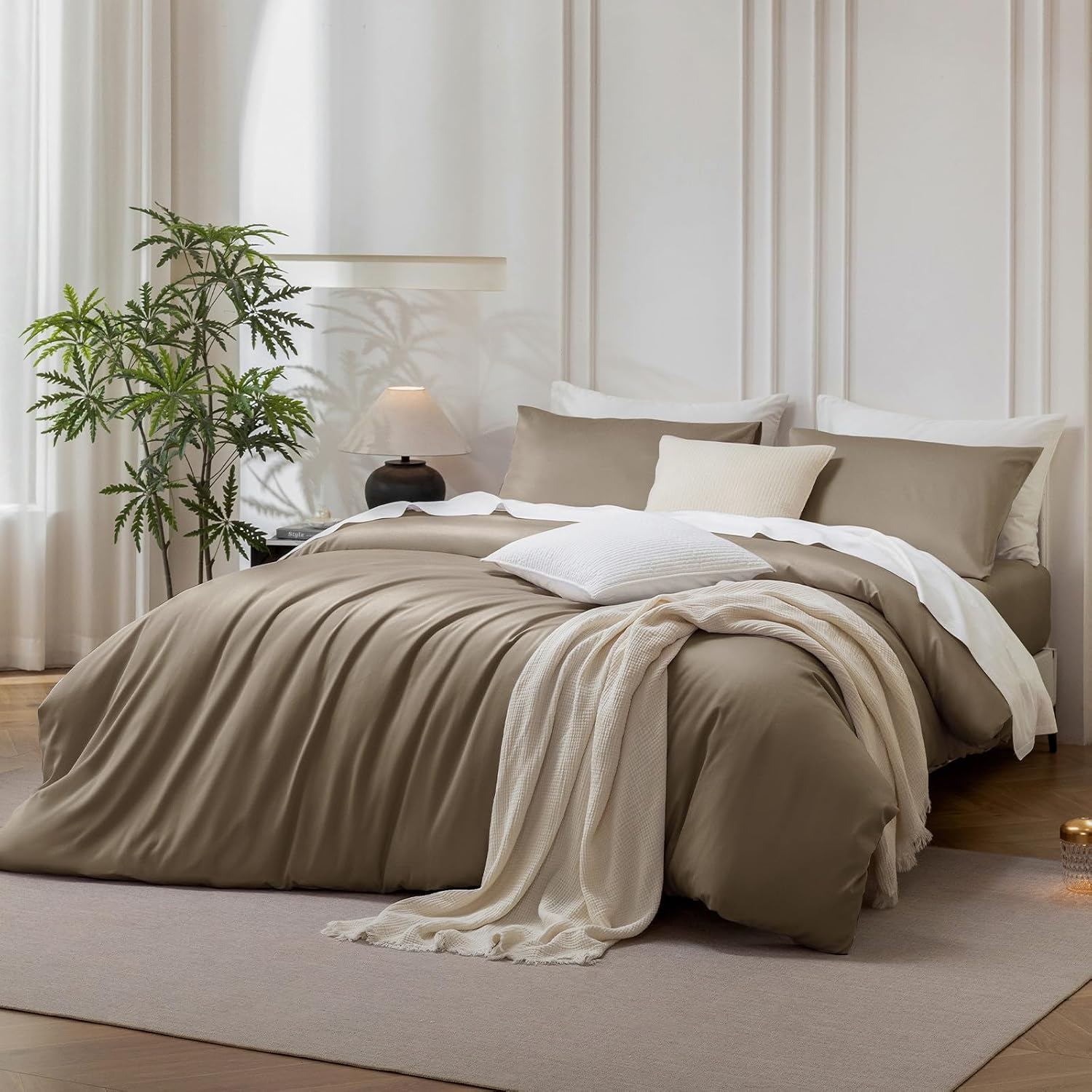 A neatly made bed in a softly lit bedroom, dressed in taupe and cream bedding with white pillows and a cream throw blanket. The room has paneled walls, a plant, a bedside lamp, and a light rug.