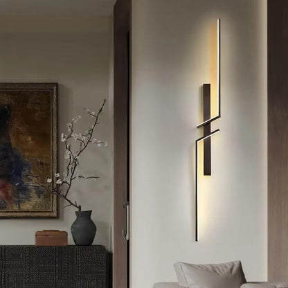 A modern wall-mounted light fixture with a matte black geometric design and warm vertical illumination, shown installed on a beige wall in a contemporary living room setting next to a sofa.