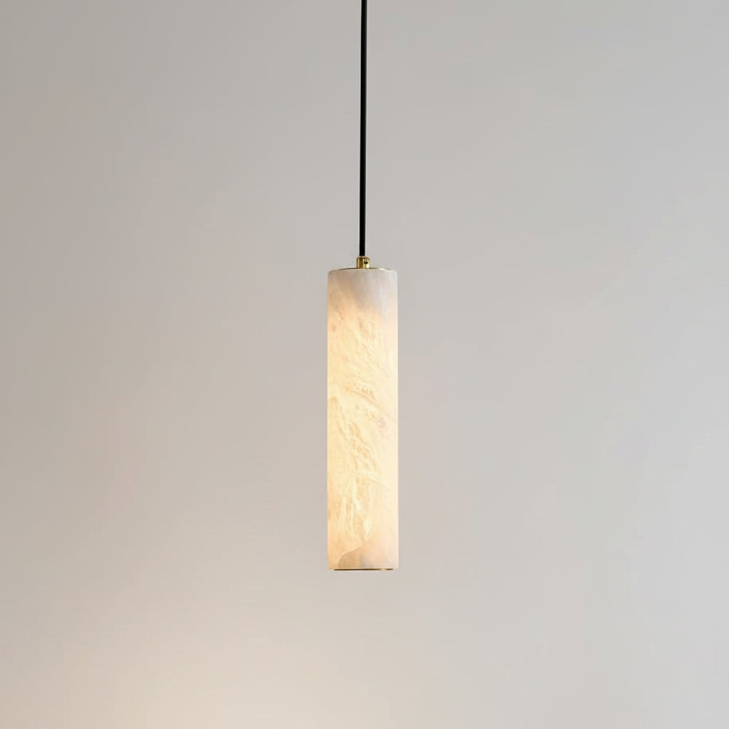 A modern cylindrical pendant light with a warm white marble-look textured shade, suspended by a black cord against a beige wall, with a framed print and wooden chair partially visible in the room setting.