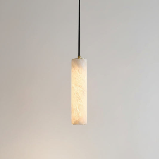 A modern cylindrical pendant light with a warm white marble-look textured shade, suspended by a black cord against a beige wall, with a framed print and wooden chair partially visible in the room setting.