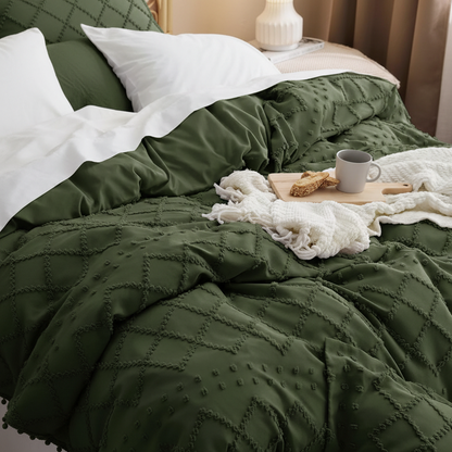 An olive green quilted comforter with pom-pom trim and matching pillow shams on a bed, styled with white pillows, a white throw blanket, and a small tray with a mug.
