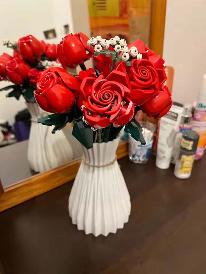 Bouquet of red roses in a white vase on a table with various items in the background.