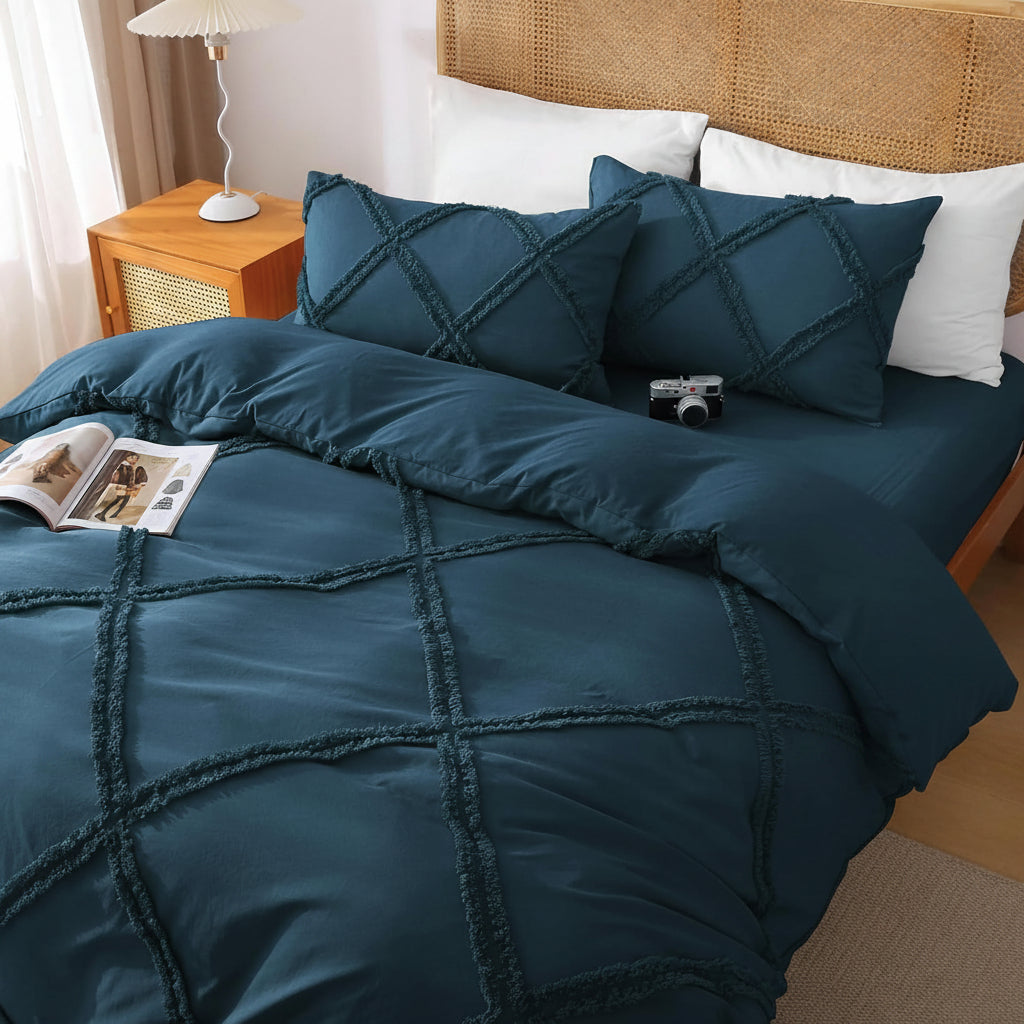 A bed dressed in a dark teal quilted bedding set with matching pillow shams, styled in a bedroom setting with a wooden headboard, bedside lamp, and an open magazine on the duvet. The text 'No Comforter' is overlaid in the bottom right corner.
