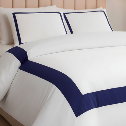 White bedding set with navy blue border trim on a bed, featuring two matching pillowcases and a duvet cover, styled with a beige tufted headboard.