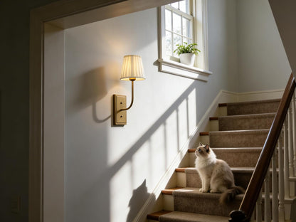 Cat sitting on a staircase in a home with a wall-mounted lamp and window in the background.