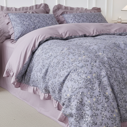 A bed dressed in a dusty lavender and gray floral print bedding set with ruffled edges, including a comforter, matching pillow shams, and a light pink bed skirt, set against a white paneled wall.
