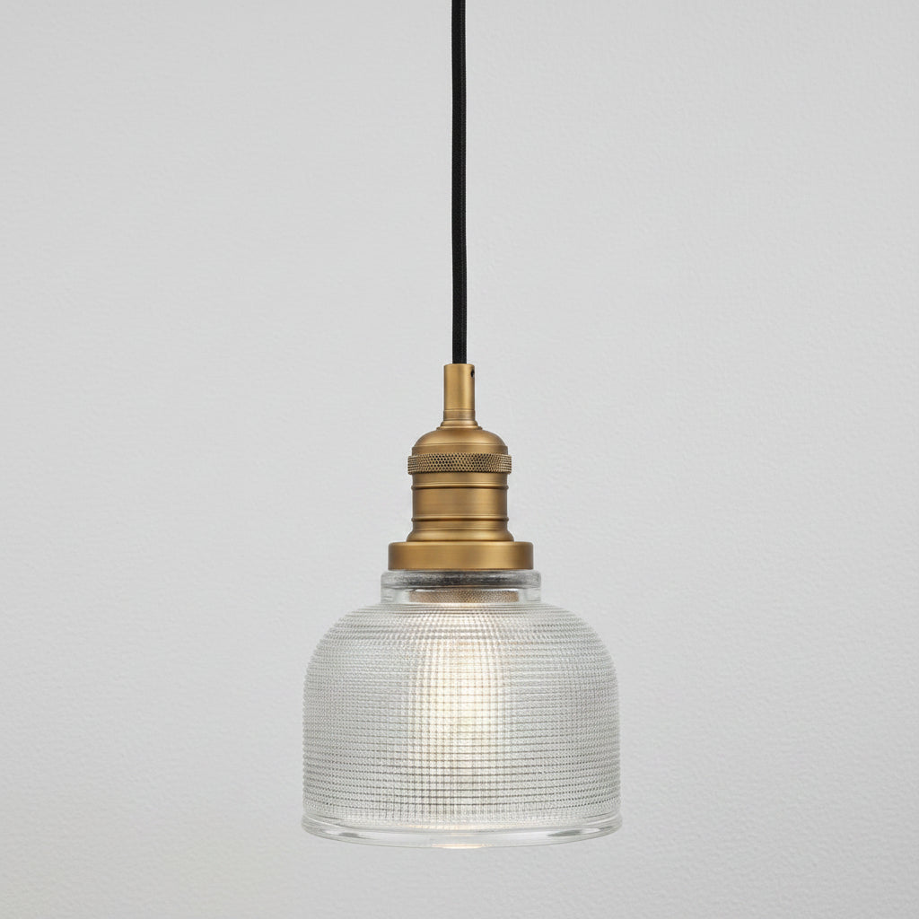 A vintage-style industrial pendant light with a clear ribbed glass dome shade, a brass metal socket, and a black hanging cord, shown against a plain white background.