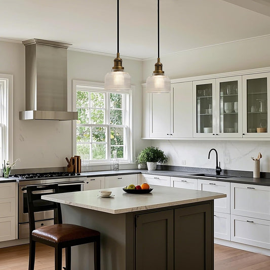 A modern kitchen interior with white shaker-style cabinets, a central island with a light stone top and dark gray base, black countertops, a stainless steel range hood, and two pendant lights hanging above the island.