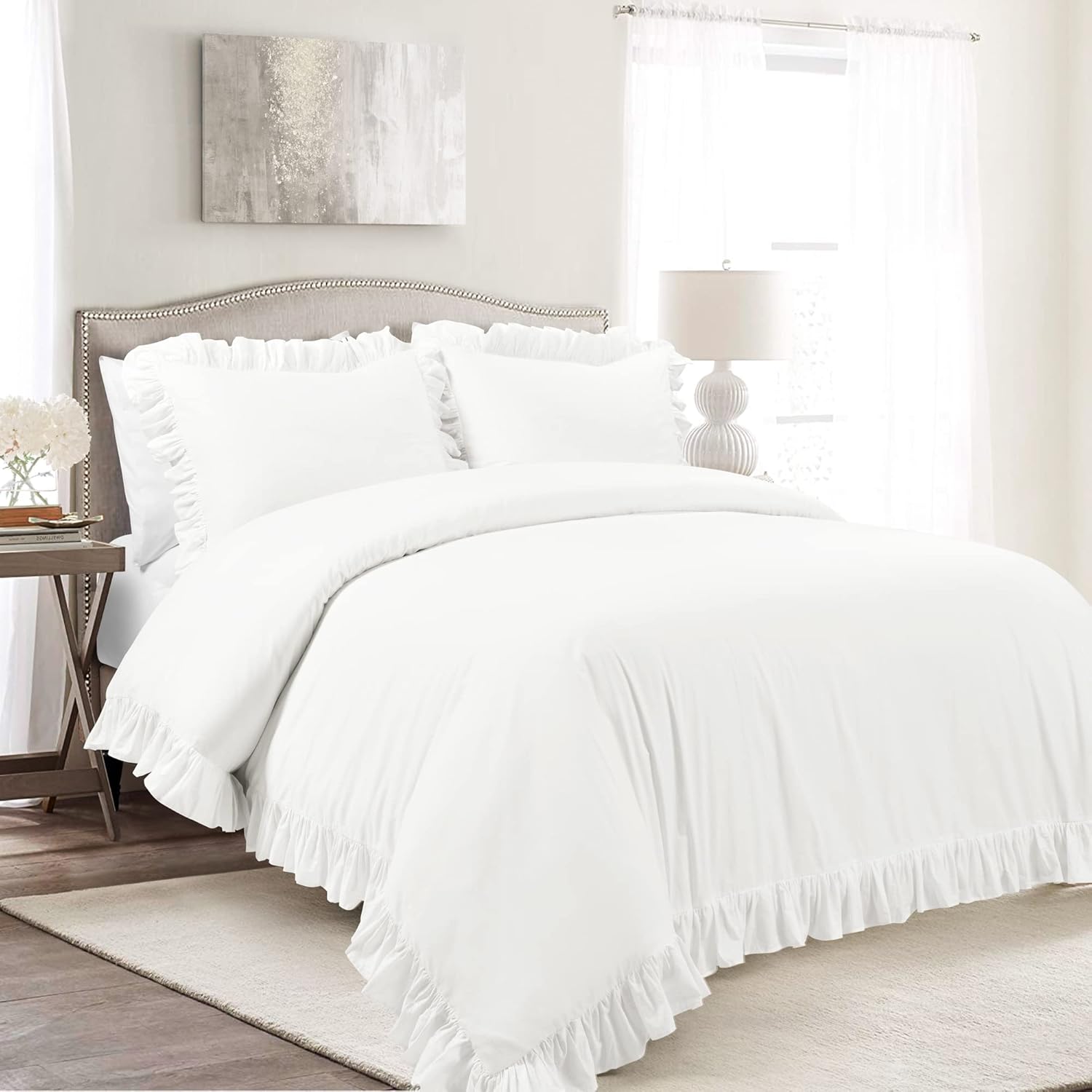 A white ruffled bedding set, including a comforter and matching pillows, styled on a bed in a bright, neutral-toned bedroom.