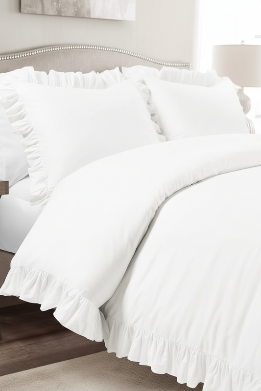 White ruffled bedding set on a bed, including a duvet cover and matching pillow shams, styled in a softly lit bedroom setting.