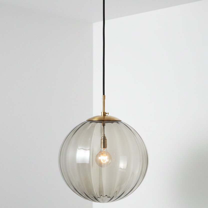 A modern pendant light fixture featuring a large, ribbed, smoky translucent glass globe shade suspended from a brushed brass ceiling canopy and cord.