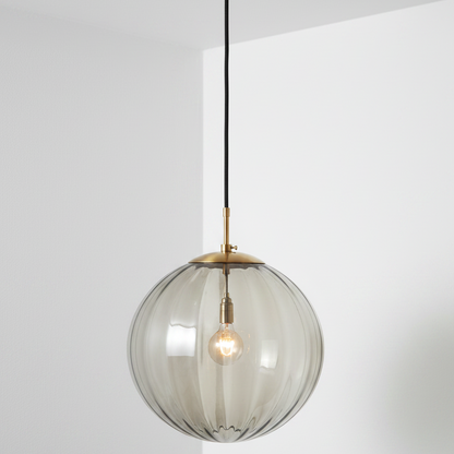 A modern pendant light fixture featuring a large, ribbed, smoky translucent glass globe shade suspended from a brushed brass ceiling canopy and cord.