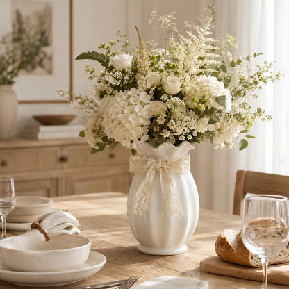 Decorative floral arrangement in a white vase on a table with tableware and bread.