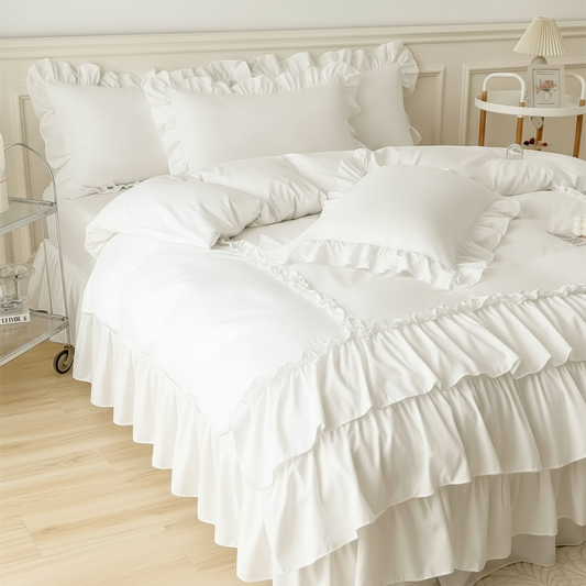 White ruffled bedding set on a bed in a bright bedroom, including a duvet cover, multiple pillows, and a ruffled bed skirt.