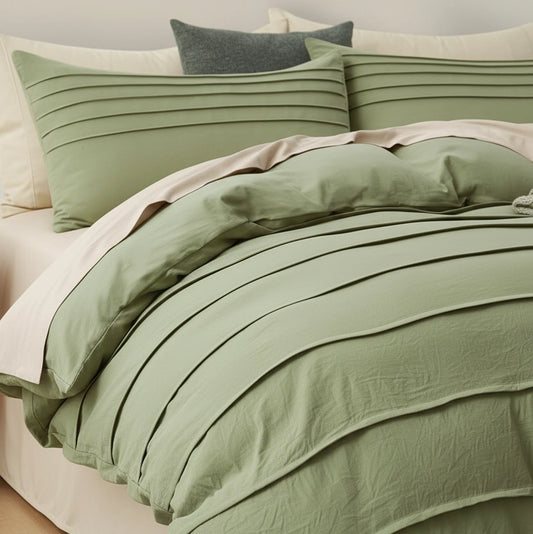 A bed dressed in a sage green bedding set with pleated textures, including a duvet cover, matching pillow shams, and a light blush pink sheet, styled with cream pillows and a dark gray accent pillow.