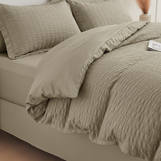 A beige textured bedding set on a bed, featuring a crinkled quilt, matching pillow shams, and a solid beige folded backsheet or duvet cover.