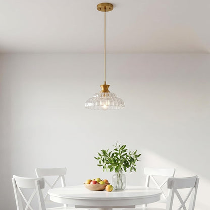 A dining room scene with a gold-finished pendant light hanging above a round white table and four white chairs. The pendant light has a clear ribbed glass shade.