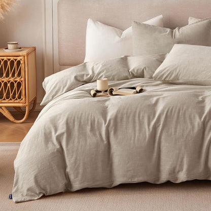A neatly made bed with light beige linen bedding and multiple white pillows, styled in a warm neutral bedroom with a woven nightstand and soft wall paneling.