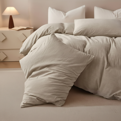 A bed dressed in light beige or oatmeal-colored bedding, including a duvet cover and multiple pillows, styled in a softly lit bedroom with a bedside lamp and nightstand.