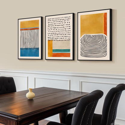 Three abstract artworks on a wall above a wooden table with chairs.