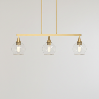 A modern three-light linear pendant chandelier with a brushed brass/gold horizontal bar, three clear glass globe shades, and visible Edison-style bulbs, shown against a textured beige wall.