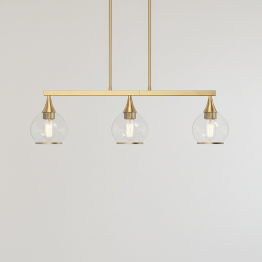 A modern three-light linear pendant chandelier with a brushed brass/gold horizontal bar, three clear glass globe shades, and visible Edison-style bulbs, shown against a textured beige wall.