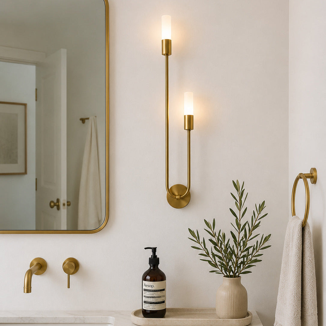 A modern bathroom scene featuring a gold wall sconce with two lit candle-style bulbs mounted on a white wall above a marble countertop with a sink, wooden vanity, mirror, soap dispenser, and a small potted plant. A gold towel ring with a white towel is visible on the right.