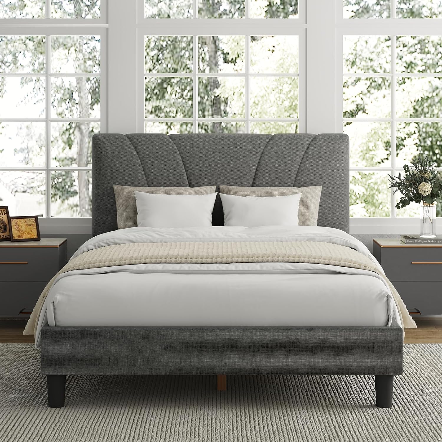 Gray upholstered bed in a bright bedroom with large windows.
