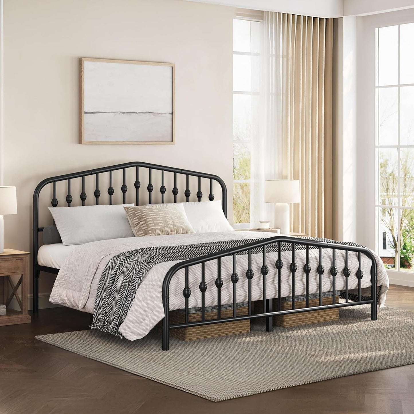 A black metal bed frame with decorative spindle-style headboard and footboard rails, shown in a styled bedroom setting with white bedding, a patterned throw blanket, a rug, and large windows with sheer curtains.