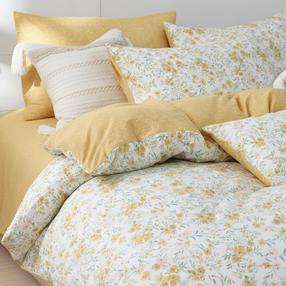 A cozy bed styled with a floral duvet cover and matching pillows in yellow and white, accented by a textured cream pillow, set against a light wood-paneled wall.