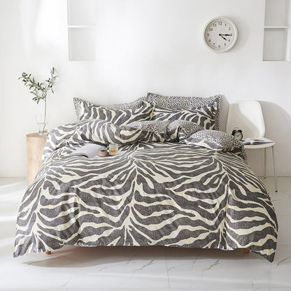 A bed dressed in a zebra-print bedding set with black, gray, and off-white stripes, styled in a bright, modern bedroom with a white chair, side table, and wall clock.