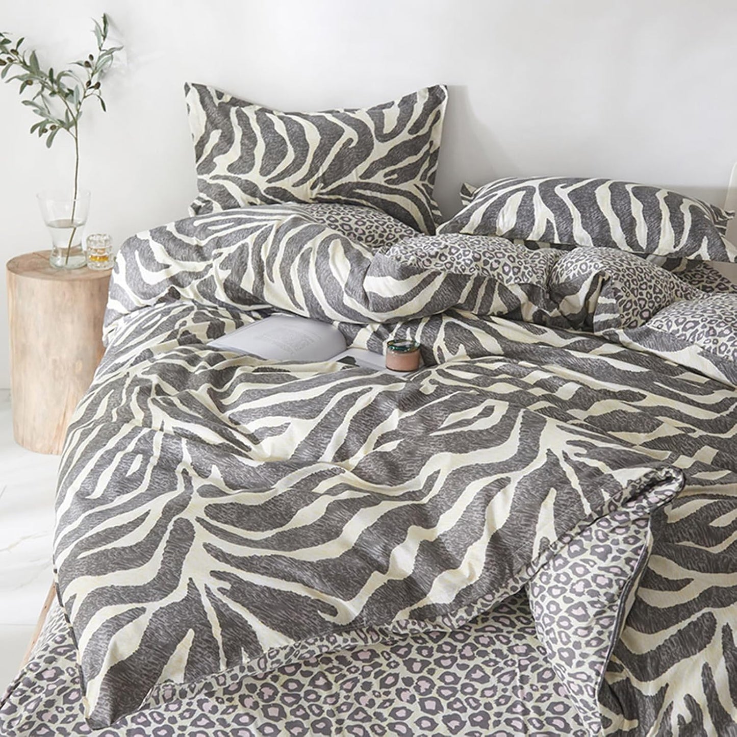 A bedding set styled on a bed, featuring a duvet cover and pillowcases with a gray and off-white zebra print, mixed with leopard print accents on the sheets and side pillows. The bed is styled in a bright, minimalist bedroom with a wooden side table holding a glass vase with greenery.