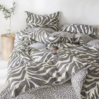 A bedding set styled on a bed, featuring a duvet cover and pillowcases with a gray and off-white zebra print, mixed with leopard print accents on the sheets and side pillows. The bed is styled in a bright, minimalist bedroom with a wooden side table holding a glass vase with greenery.