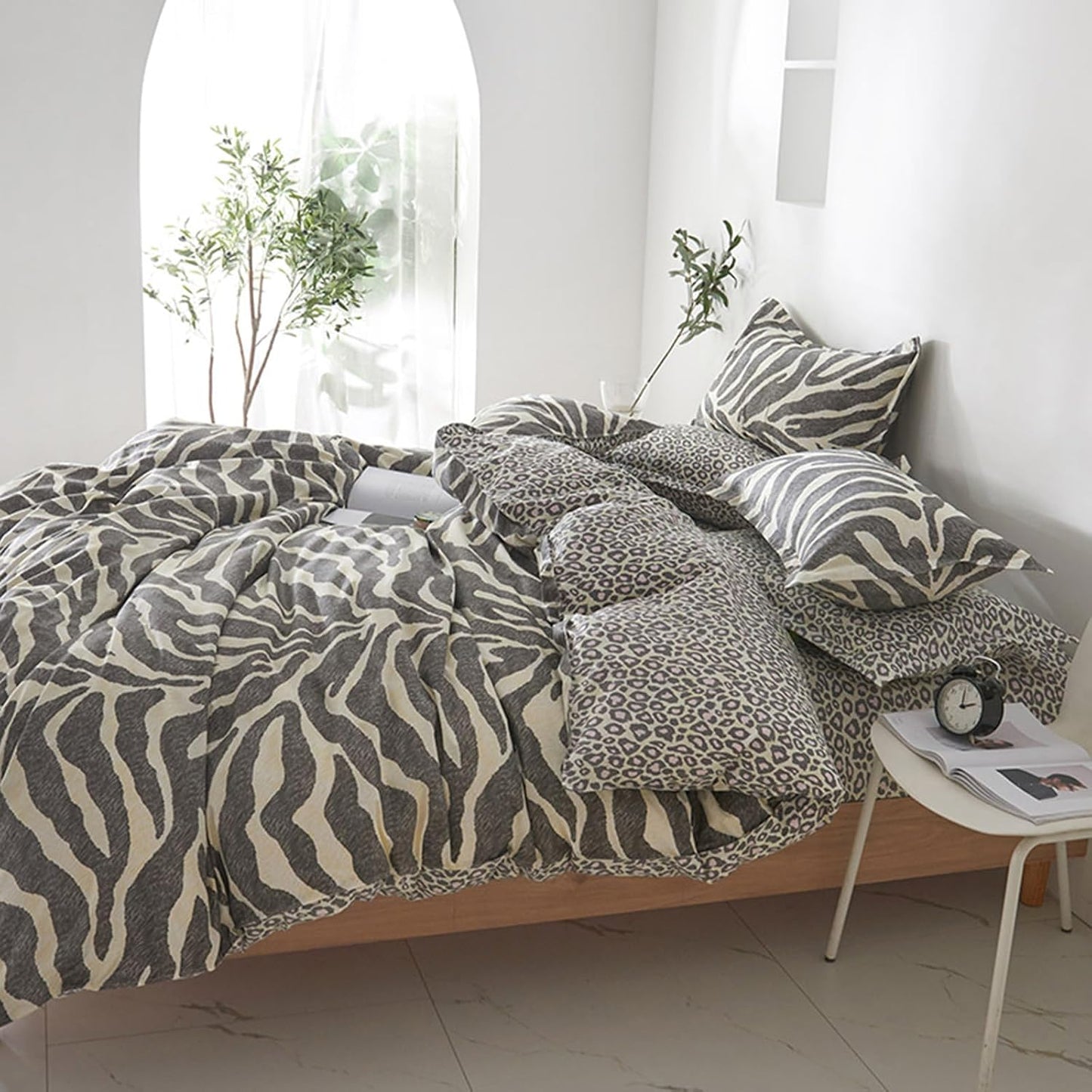 A bed dressed in a zebra-print and leopard-print bedding set, styled in a bright, modern bedroom with white walls, a small side table, and potted plants.