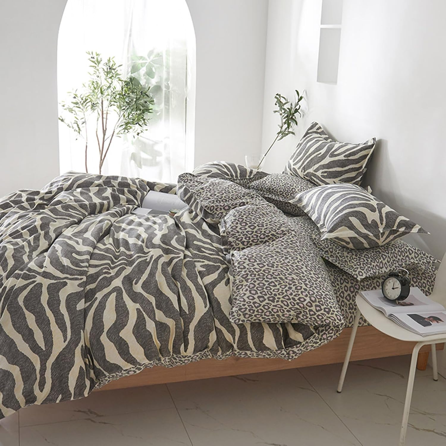 A bed dressed in a zebra-print and leopard-print bedding set, styled in a bright, modern bedroom with white walls, a small side table, and potted plants.