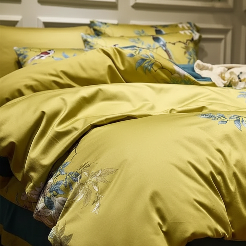 A close-up of a bed dressed in a yellow-green satin bedding set with blue and white floral embroidery, set against a warm wood-paneled wall and wooden floor.